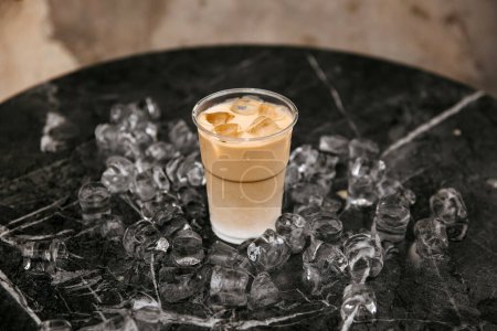 Photo for An iced coffee in a plastic cup surrounded by scattered ice cubes on a marble surface. - Royalty Free Image