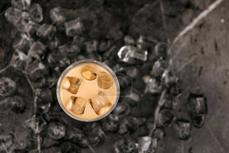 Foto de Una vista de arriba hacia abajo de un latte helado en un vaso sobre una superficie oscura y texturizada con cubitos de hielo dispersos alrededor. - Imagen libre de derechos
