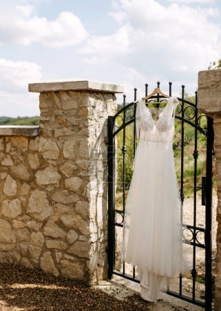 Foto de Vestido de novia blanco colgado en una puerta de hierro forjado negro con un pilar de piedra y paisaje rural en el fondo. - Imagen libre de derechos