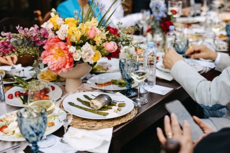 Téléchargez les photos : Cadre de table festive avec pièce maîtresse florale colorée, repas des invités, plat de poisson, asperges, vin, eau et atmosphère vibrante. - en image libre de droit