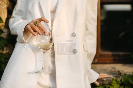 Téléchargez les photos : Femme élégamment habillée tenant un verre de vin blanc, la lumière du soleil coulant une douce lueur. - en image libre de droit