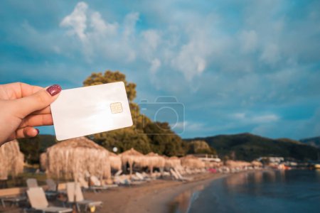 Weiße Bankkarte in der Hand der Frau auf dem Hintergrund des Strandes mit Sonnenliegen und Sonnenschirmen in Moraitika, Korfu, Griechenland. Das Konzept der Bezahlung für Entspannung und unbegrenzte Möglichkeiten. Hochwertiges Foto