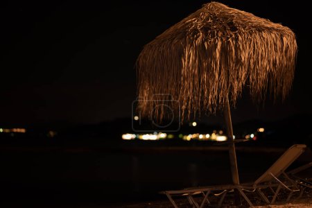 Téléchargez les photos : Tournage nocturne de chaises longues de plage et parapluie en feuilles de palmier balancent dans le vent sur fond de mer ionienne et de ciel noir Moraitika, Corfou, Grèce. Photo de haute qualité - en image libre de droit