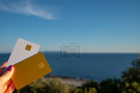 Foto de Golden and White Bank Card In Woman Hand On Background Of Scenic View From Arkoudilas Viewpoint, Mountains, Jonian Sea Corfu, Greece (en inglés). El concepto de pago para relajarse, posibilidades ilimitadas. Alto. - Imagen libre de derechos