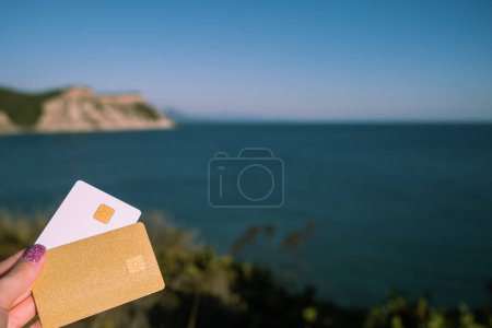 Golden and White Bank Card In Woman Hand On Background Of Scenic View From Arkoudilas Viewpoint, Mountains, Ionian Sea Corfu, Greece. The Concept Of Payment For Relax, Unlimited Possibilities. High