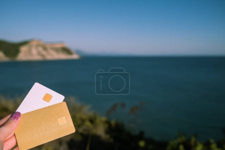 Goldene und weiße Bankkarte in Frauenhand vor dem malerischen Hintergrund des Aussichtspunktes Arkoudilas, Berge, Ionisches Meer Korfu, Griechenland. Das Konzept der Bezahlung für Entspannung, unbegrenzte Möglichkeiten. Hoch