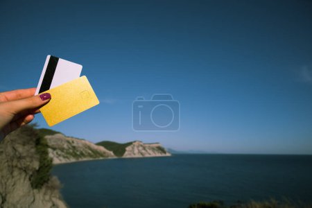 Foto de Golden and White Bank Card In Woman Hand On Background Of Scenic View From Arkoudilas Viewpoint, Mountains, Jonian Sea Corfu, Greece (en inglés). El concepto de pago para relajarse, posibilidades ilimitadas. Alto. - Imagen libre de derechos