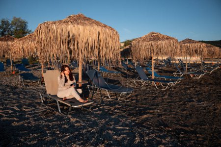 Téléchargez les photos : La jeune femme au lever du soleil s'assoit sur une chaise de plage sous un parapluie et allume son casque pour profiter de la musique et de la vue pittoresque. Concept de détente et d'immersion dans vos pensées. . Haute qualité - en image libre de droit
