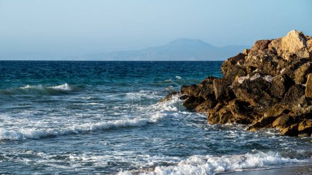 Téléchargez les photos : Paysage envoûtant avec la mer dans laquelle les vagues font rage et s'écrasent contre les brise-lames en pierre, avec des montagnes visibles au loin. . Photo de haute qualité - en image libre de droit