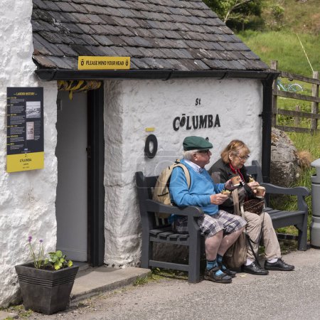 Photo for Isle of Iona, Scotland, UK. 6 June 2023. Tourists stop for lunch seated on a wooden bench outside a larder selling food and drinks on the small island of Iona. - Royalty Free Image