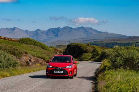 Photo for Isle of Skye,  Scotland, UK.  5 June 2023. Small red car and scenic countryside view across to the Cuillin Hills with a blue sky. Isle of Skye, UK. - Royalty Free Image