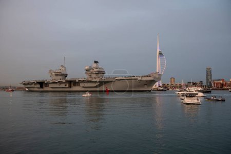 Photo for Portsmouth, England, UK. 8 September 2023.  HMS Queen Elizabeth underway leaving Portsmouth dockyard at dusk with an escort of tugs and security vessels. Naval personnel line the decks. - Royalty Free Image