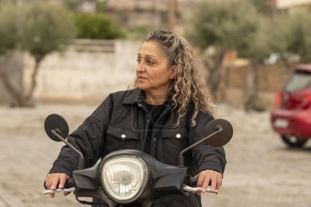 Photo for Malia, Crete, Greece. 03.10.2023. Cretan woman with curly hair and wearing a blck jacket riding a motor scooter. - Royalty Free Image