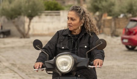 Photo for Malia, Crete, Greece. 03.10.2023. Cretan woman with curly hair and wearing a blck jacket riding a motor scooter. - Royalty Free Image