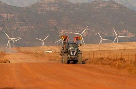 Téléchargez les photos : Gouda, Western Cape, Afrique du Sud. 24 avril 2024. Fin de journée et un tracteur agricole fait route sur un chemin de terre rouge avec un fond de parc éolien Gouda et les montagnes. - en image libre de droit