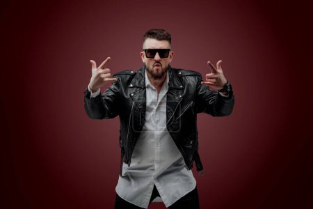 Téléchargez les photos : The man is dressed in a Ukrainian shirt; a man in a leather jacket poses on a red background; shows different facial expressions and gestures; poses in sunglasses in the studio; charismatic portrait of a man with a beard - en image libre de droit