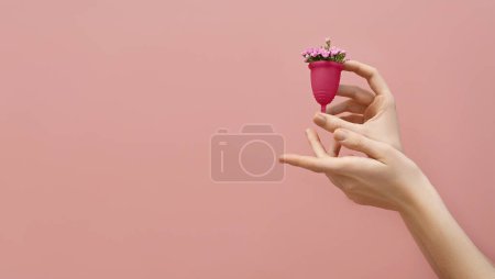 Photo for Female hand holding menstrual cup filled with flowers  on pink background, woman wellbeing concept - Royalty Free Image