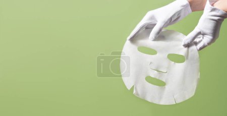 Photo for Female gloved hands holding white facial mask sheet on green background. Beauty skincare concept - Royalty Free Image