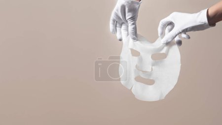 Photo for Female gloved hands holding white facial mask sheet on beige background. Beauty skincare concept - Royalty Free Image