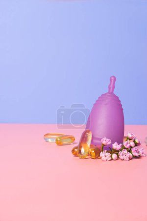 Female menstual cup, flowers and oil capsules on pink table. Woman well-being concept