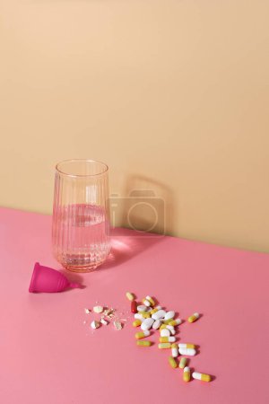 Photo for Glass of water, menstual cup and handful of pills on a pink table . Woman well-being concept - Royalty Free Image
