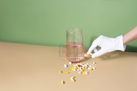 Foto de Guante de mano femenina sosteniendo una píldora. vaso de agua y un puñado de pastillas están en una mesa. Concepto de bienestar - Imagen libre de derechos