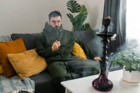 Téléchargez les photos : Bearded man is smoking hookah at home and blowing cloud of smoke - chill time and resting - en image libre de droit