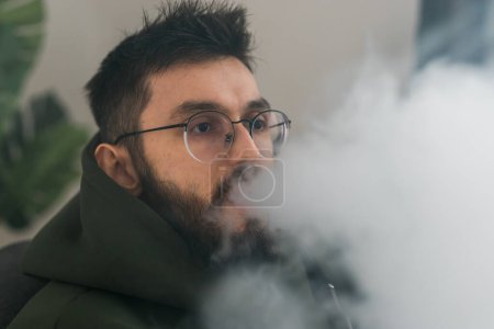 Téléchargez les photos : Bearded man is smoking hookah at home and blowing cloud of smoke - chill time and resting - en image libre de droit