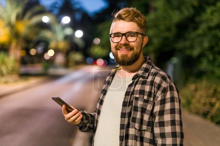 Bearded man wearing eyeglasses is holding scrolling texting in his cellphone at night street. Guy calls for a taxi in an app in evening city