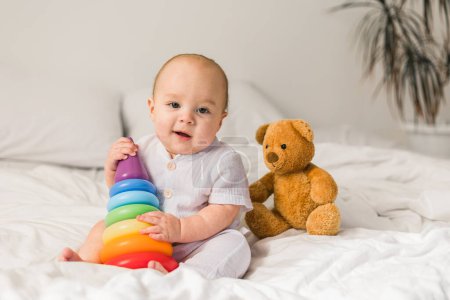 Foto de Lindo bebé jugando con colorido arco iris juguete pirámide y oso de peluche sentado en la cama en el dormitorio blanco soleado. Juguetes para niños pequeños. Niño con juguete educativo - Imagen libre de derechos