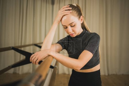 Photo for Upset ballerina in training suit stands near ballet barre and looks at the mirror in dance studio. Woman prepares for performance. Ballet dancer doing gymnastic exercises. Classical ballet school. - Royalty Free Image
