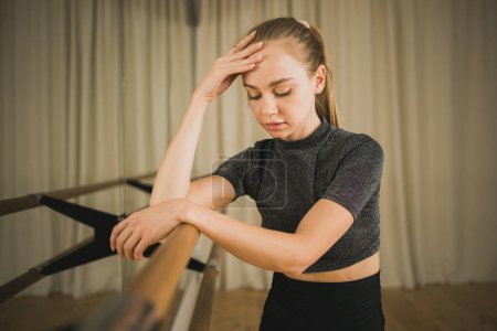 Photo for Upset ballerina in training suit stands near ballet barre and looks at the mirror in dance studio. Woman prepares for performance. Ballet dancer doing gymnastic exercises. Classical ballet school. - Royalty Free Image