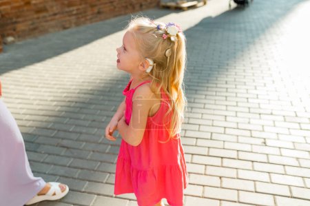 Niña con audífono implante coclear en vestido rosa que se divierte en la calle de verano. Ayuda para el tratamiento de la sordera y la pérdida auditiva en seres humanos. Copiar espacio.