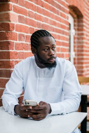 Téléchargez les photos : Afro-Américain hipster gars en vêtements culturels et ethniques dashiki bavarder sur smartphone à l'extérieur, textos avec des amis sur les réseaux sociaux. Concept Millénaire et Internet. - en image libre de droit