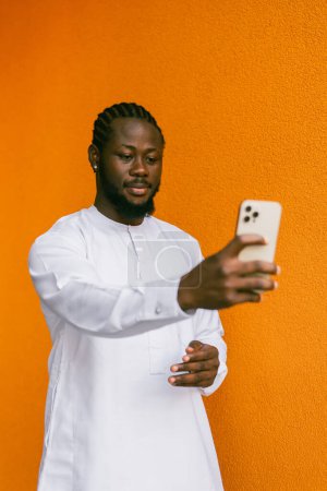Téléchargez les photos : Heureux jeune homme afro-américain en vêtements ethniques dashiki prendre selfie sur fond de mur de briques. Étudiants et jeunes de la génération millénaire - en image libre de droit