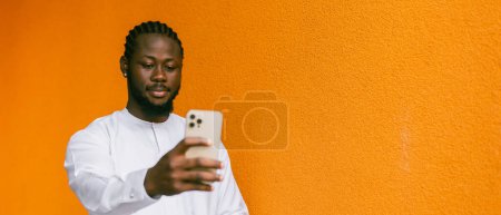 Téléchargez les photos : Bannière Heureux jeune homme afro-américain en vêtements ethniques dashiki prendre selfie sur fond mural orange copie espace. Étudiants et jeunes de la génération millénaire - en image libre de droit