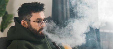 Bearded millennial or gen z man smoking hookah while relaxing on sofa at home - chill time and resting