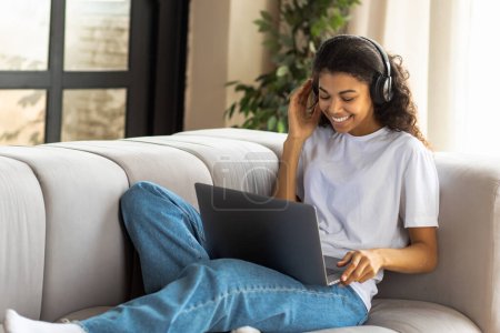 Photo for Video call, virtual chat concept. Young African American hipster woman in earphone with Afro hair using a laptop talking to webcam with friend online lying on the couch at home - Royalty Free Image