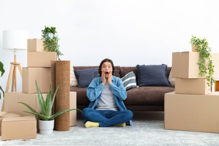 Photo for Moving to a new house rental housing. Happy young caucasian woman sitting on the floor with cardboard boxes in her new home with a surprised expression from profitable home loan looking at the camera - Royalty Free Image