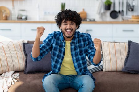 Photo for Portrait of happy Indian man overjoyed with good news. Smiling male with curly hair showing yes gesture, sitting on couch at home - Royalty Free Image