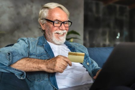 Photo for Senior smiling and looking at the laptop shopping online with credit card. Elderly person place an order for purchase of goods online sitting on the couch at home - Royalty Free Image