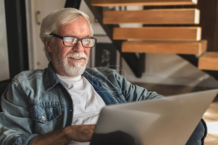 Photo for Elderly man with glasses with gray hair and beard sits at home on the sofa works or browses social networks on laptop. Happy carefree pension - Royalty Free Image