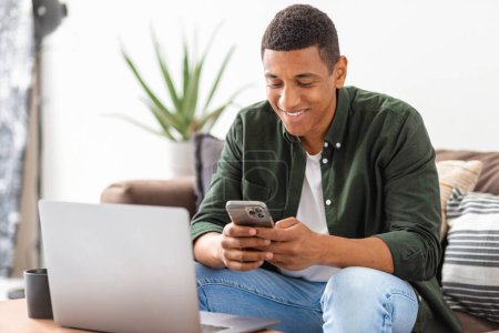 Photo for Successful man entrepreneur or freelancer sitting on sofa in home office chatting using smartphone. Young student male working, using mobile phone and laptop, smiling - Royalty Free Image