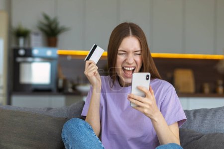 Photo for Overjoyed Caucasian woman with open mouth holding mobile phone and credit card sitting at home on the couch. Online shopping, technology concept - Royalty Free Image