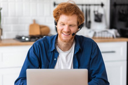 Photo for Remote online work. Happy caucasian man talking online using headset and laptop. Freelancer working remotely online sitting at home at the table - Royalty Free Image