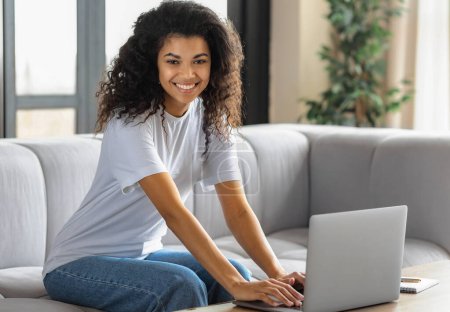 Photo for Young African American woman looking at camera and smiles. Female student or freelancer working or watching webinar using laptop sitting on at home. Online learning, distance education - Royalty Free Image