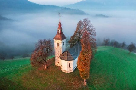 Foto de Vista superior de la pequeña iglesia hermosa en una montaña en Eslovenia al amanecer o al atardecer. Hermoso paisaje matutino de otoño, tiro aéreo con dron - Imagen libre de derechos
