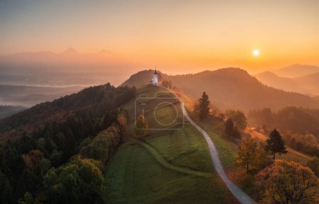 Foto de Vista aérea del dron de la pequeña iglesia hermosa en la cima de una montaña en Eslovenia al amanecer. Hermoso paisaje de otoño mañana - Imagen libre de derechos