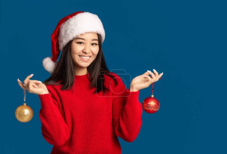 Photo for Happy woman holds in hands christmas balls. Beautiful Asian woman in santa claus hat on blue background, Looking at the camera and smiles. Xmas concept - Royalty Free Image