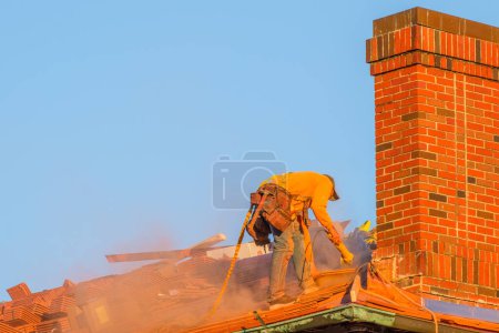 Téléchargez les photos : New Orleans, LA, USA - January 4, 2023: Roofer cutting roofing tiles with a power saw and putting orange dust in the air next to a chimney during the late afternoon - en image libre de droit